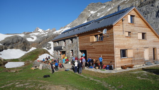 Claridenhuette SAC Die Claridenhuette in der Sonne. Vor der Huette befinden sich circa 20 Personen mit Rucksaecken.